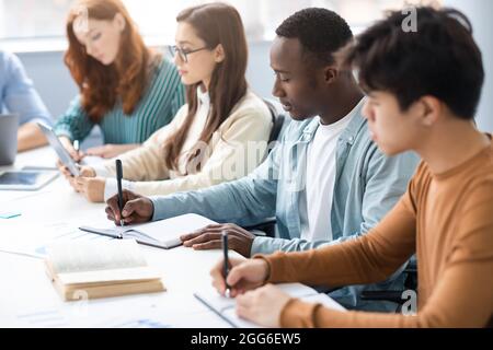 Diverse Students Sitting At Desk In Line Taking Notes Stock Photo