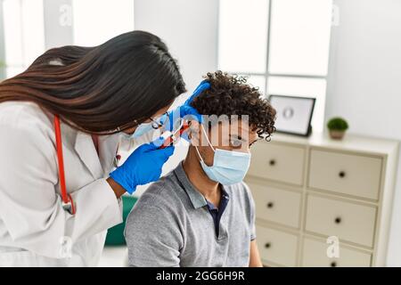 Young latin doctor woman auscultating the ear of man using otoscope at examination room. Stock Photo