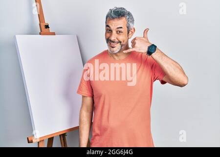 Handsome middle age man with grey hair standing by painter easel stand smiling doing phone gesture with hand and fingers like talking on the telephone Stock Photo