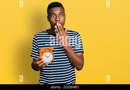 Young african american man holding alarm clock covering mouth with hand, shocked and afraid for mistake. surprised expression Stock Photo