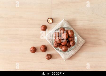 Raw macadamia nuts in a transparent glass bowl and scattered on wooden background. Minimal composition. Stock Photo