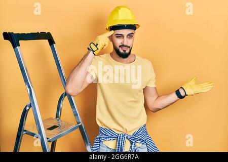Handsome man with beard by construction stairs wearing hardhat confused and annoyed with open palm showing copy space and pointing finger to forehead. Stock Photo