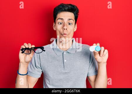 Young hispanic man holding glasses and contact lenses making fish face with mouth and squinting eyes, crazy and comical. Stock Photo