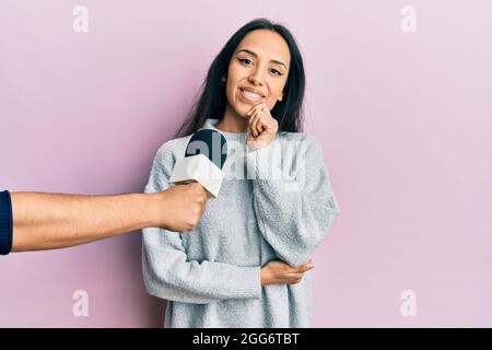 Young hispanic girl being interviewed by reporter holding microphone smiling looking confident at the camera with crossed arms and hand on chin. think Stock Photo