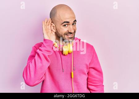Young bald man wearing gym clothes and using headphones smiling with hand over ear listening an hearing to rumor or gossip. deafness concept. Stock Photo