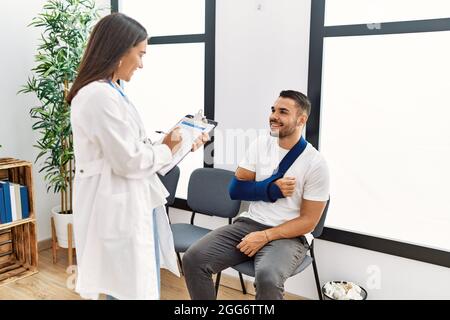 Youg latin injuried man wearing sling talking with the doctor at clinic waiting room. Stock Photo