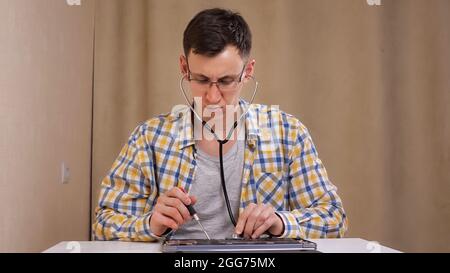 man with glasses examines a disassembled laptop and listens with a phonendoscope Stock Photo