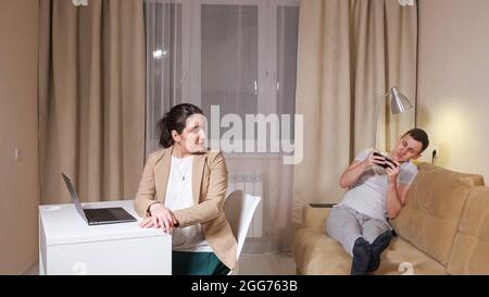 Concentrated brunette types on grey laptop sitting on chair at table and husband lying on sofa and playing game Stock Photo