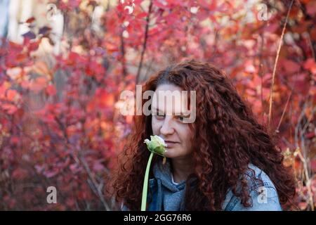 woman with flower close up. woman with long curvy brown hair smell white flower in autumn forest with red bush on background Stock Photo