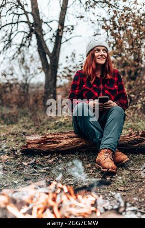 Traveler girl in hipster clothes sits on a log enjoying, relaxing in the forest near the fire by camping. Woman using smartphone in nature Stock Photo