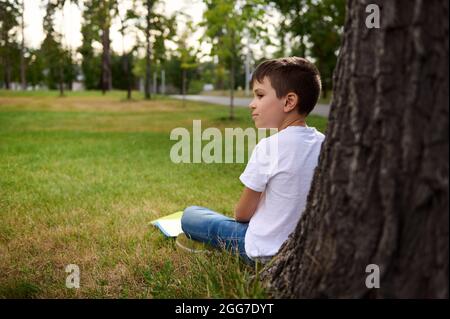 Side view of distracted schoolboy enjoying recreation after school , sitting on the grass of the park, leaning against a tree, doing his homework and Stock Photo