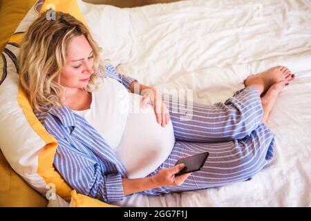 Pregnant woman in blue striped pajamas looking in smartphone in bedroom in the morning after waking up. A woman waiting for a child in the ninth month Stock Photo