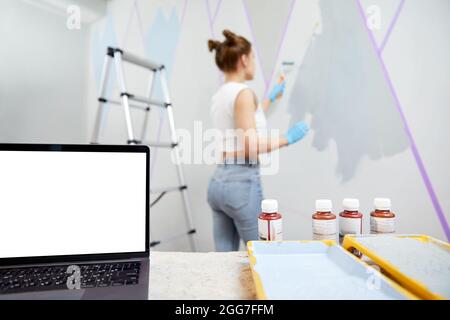 Young woman painting wall with paint roller and using masking tape. Laptop standing on table in front with white copy space mockup Stock Photo