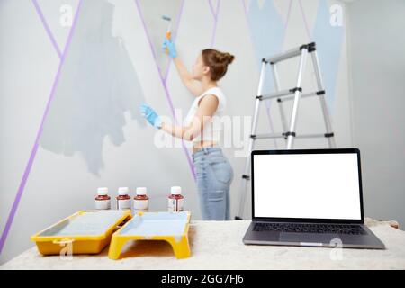 Young woman painting wall with paint roller and using masking tape. Laptop standing on table in front with white copy space mockup Stock Photo