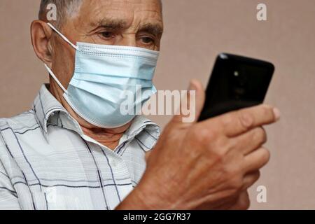 Elderly man in protective face mask using mobile phone, looking on smartphone display. Concept of online communication in retirement, sms, safety duri Stock Photo