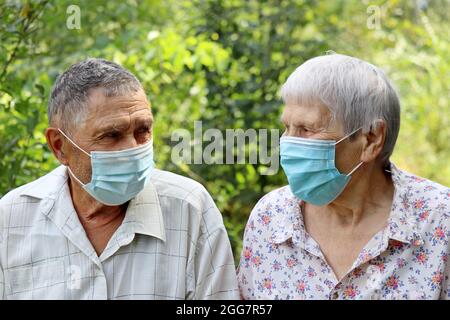 Elderly couple in protective face masks looking at each other on nature background. Safety during coronavirus pandemic, life in retirement Stock Photo