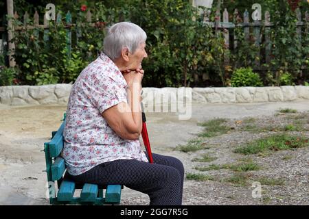 Elderly woman sitting with walking cane on a bench in a yard. Healthy lifestyle in old age, life in retirement Stock Photo