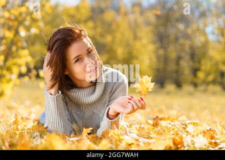 Young woman feels no problem as little girl concept. A lady in gray knitted sweater lies on fallen yellow dry autumn leaves on sunny day. The head is in the foreground. Rest in the park and forest. Stock Photo