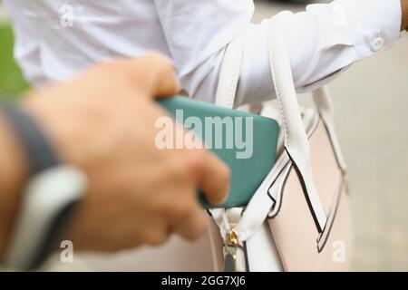 Thiefs hand steals a wallet from a womans bag, back view. A criminal steals a smartphone from a careless woman Stock Photo