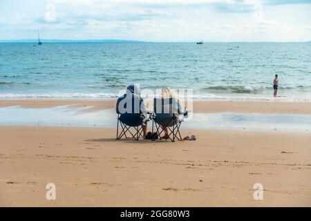 A couple of people sit alone in folding chairs close to the water's edge on Tenby North Beach in Wales, UK. Stock Photo