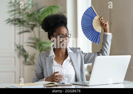 Exhausted businesswoman tired of heat waving paper fan for fresh air at workplace work on laptop Stock Photo