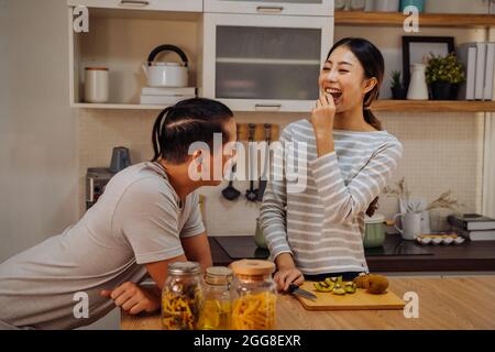 Young married couple cooking together in kitchen at home. Young woman feeding her man with kiwi and fruits indoors Stock Photo