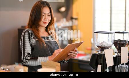 Pretty young barista looking at drinks menu, holding menu, taking order from customer, coffee shop, cafe Stock Photo