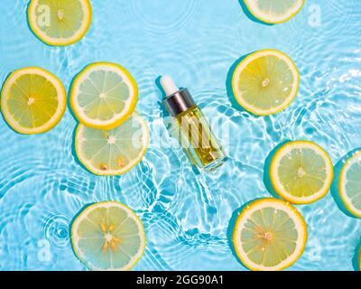 Luxury cosmetic oil or serum in glass bottle with pipette in clean transparent water with lemon slices over blue background. Water splashing on water surface in sunlight. Vertical flatlay copy space Stock Photo
