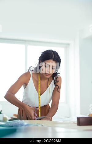 Pretty young seamstress cutting expensive fabric for sharp scissor when working on order for customer in her workshop Stock Photo