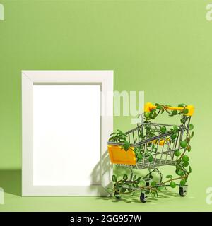 Shopping cart entwined with plants next to empty mock up frame on green background. Conscious consumption Stock Photo