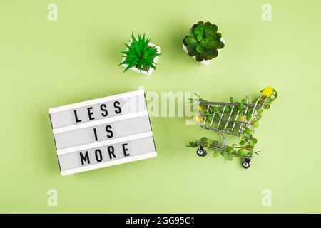 Less is more - written on lightbox next to Shopping cart entwined with plants and succulents in pots on green background. Stock Photo