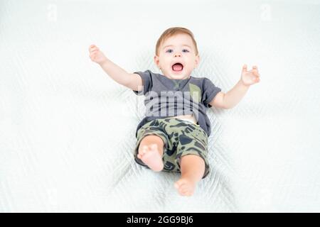 Baby boy in a khaki suit playing and shouting on white bed. Portrait of adorable toddler on white background, copy space. Stock Photo