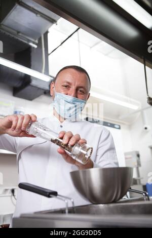 Vertical portrait of a chef in medical face mask salting prepared meal at restaurant kitchen Stock Photo