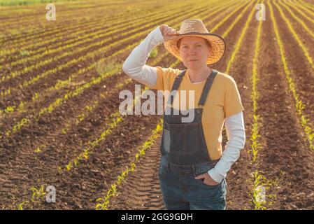 Portrait of female corn farmer in cultivated maize field wearing straw hat and jeans bib overalls and standing among young crop seedlings Stock Photo