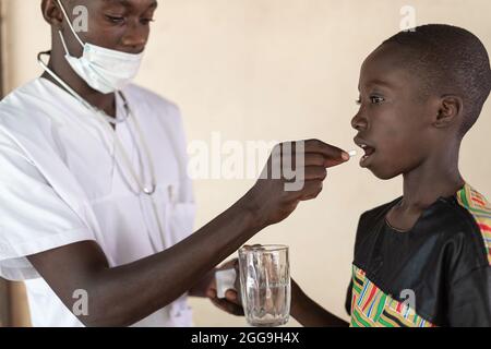 Health professional with protective mask and stethoscope giving a pill to a smal black boy holding a glass of water; child health care concept Stock Photo