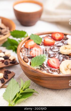 Chocolate cornflakes with milk and strawberry in wooden bowl on white wooden background and linen textile. Side view, close up, selective focus. Stock Photo