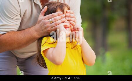 Happy father surprising his daughter in yellow t-shirt closing her eyes by hands standing behind her, playing a game guess who. Daddy day concept. Stock Photo