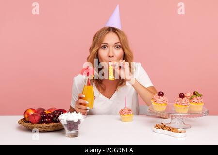 Blonde woman celebrating birthday, blowing horn, drinking cocktail, looking at camera, wearing white T-shirt and party cone, enjoying cakes. Indoor st
