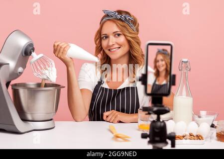 Smiling young adult blonde food blogger showing process of making cakes online, shows pastry cone for decorating to smart phone on tripod. Indoor stud Stock Photo