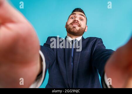 Portrait of positive bearded handsome young adult man wearing official style suit taking selfie, looking at camera with funny smile , POV. Indoor stud Stock Photo