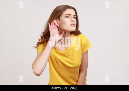 What? I can't hear! Portrait of girl in yellow T-shirt holding hand near ear and listening carefully, having hearing problems, deafness in communicati Stock Photo