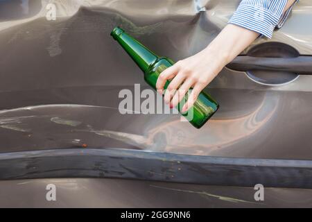Unknown female hand holding bottle with beer white sitting behind wheel of broken car, road accident due to drunk driving, damaged side part of automo Stock Photo