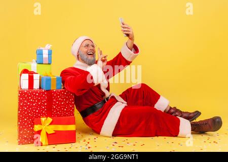 Side view of elderly man with gray beard wearing santa claus costume sitting on floor near gifts, having video call, showing v sign to followers. Indo Stock Photo