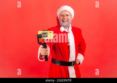 Attractive elderly man with gray beard wearing santa claus costume holding steadicam, broadcasting livestream, blogger talking with followers. Indoor Stock Photo