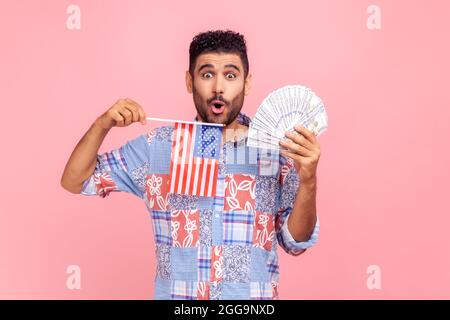 Astonished attractive man with beard and dark hair holding USA flag and dollars bills, looking at camera with open mouth, expressing shock. Indoor stu Stock Photo