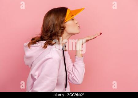 Profile of curly haired teenage girl in hoodie and yellow sun visor with plastic shield sending air kiss, flirt and affection. Indoor studio shot isol Stock Photo