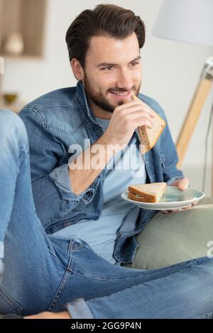 man eating a sandwich sat on the sofa Stock Photo
