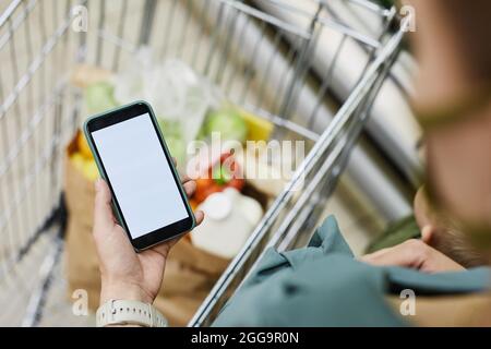 Over shoulder view of unrecognizable woman standing at shopping cart and using mobile app while buying food Stock Photo