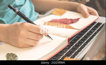 Diary book with autumn leaves and hand with pencil close-up view. Autumn hygge background Stock Photo