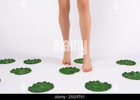 Cropped photo of slim young woman legs with red pedicure walking on tiptoes among lily leaves from plastic after epilation procedure. Stock Photo